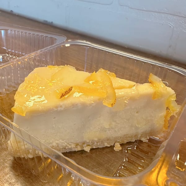 A slice of cheesecake with candied lemon peel topping in a clear plastic container.